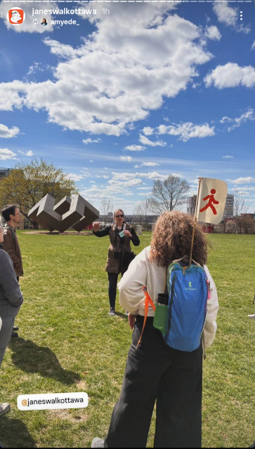 That's me gesturing about something while clasping cue cards. Standing on a gray field with a large geometric sculpture behind me and a blue sky with perfectly fluffy clouds. The Jane's walk volunteer holding the Jane's walk flag is in the foreground.