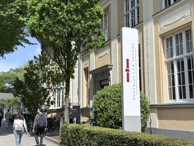 Bild einer Straßenszene mit dem Schild der Bucerius Law School. Der Hintergrund umfasst ein Gebäude mit großen Fenstern und Grün, flankiert von Bäumen. Zwei Personen gehen den Weg entlang, Fahrräder sind in der Nähe geparkt. Klarer Himmel deutet auf einen sonnigen Tag hin.
