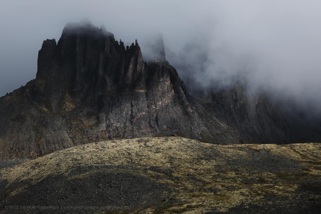A massive dark cliff face with a crown of jagged, upward-thrusting rock spires fills the upper portion of the frame, its upper ridgeline partially swallowed by low cloud moving across from the right. A rounded, lichen-covered scree ridge in the foreground is lit by a shaft of pale light, its yellow-green tones contrasting sharply with the dark, brooding rock face rising steeply behind it.