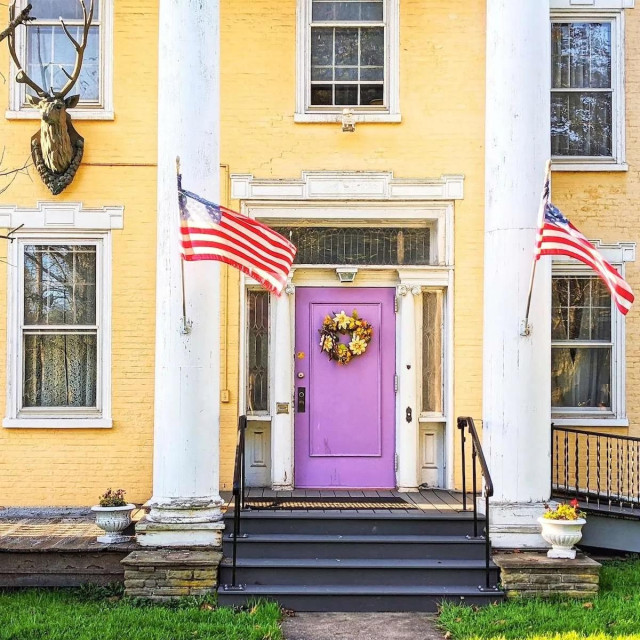 A bright purple front door adorned with a floral wreath, centered on a pale yellow brick house. Two large white columns frame the porch, each flying an American flag, with a mounted stag head visible on the wall to the left.