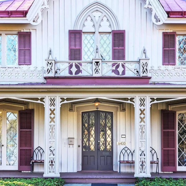 A pair of dark grey-purple doors with diamond-paned glass, situated on a white Gothic Revival house with decorative "gingerbread" trim. The entrance is flanked by purple shutters and white latticework porch supports.