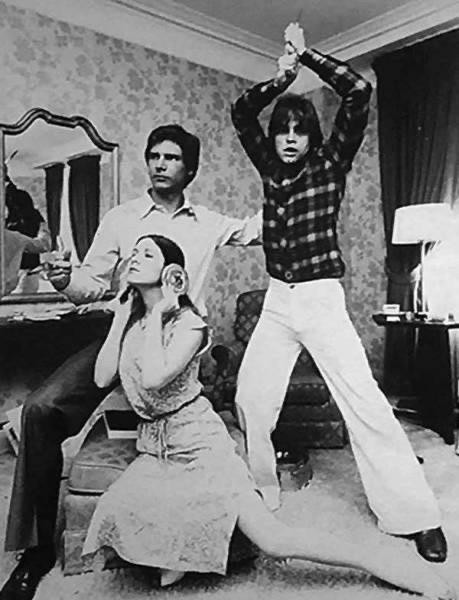 B&W photo (1970s) of Mark Hamill, Carrie Fisher & Harrison Ford, in street clothes, recreating the original "Star Wars" poster in a hotel room. Fisher crouches in front holding cinnamon rolls to her ears in place of the iconic hairdo.