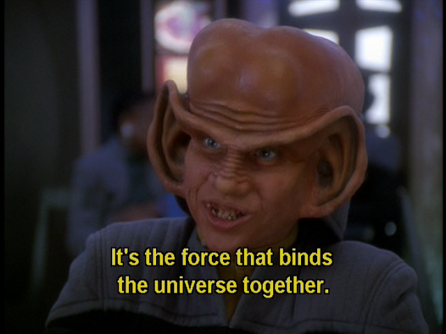 Star Trek Deep Space Nine scene. Nog is pictured in Quark's Bar up close and in Starfleet uniform. He's a Ferengi, and has giant ear lobes significantly bigger than a humans and the top curve of the ear extends and connects across like a unibrow. Also from the front, his bald head is kind of shaped like a beluga whale. He's explaining something here with enthusiasm. Closed caption reads, "It's the force that binds the universe together."