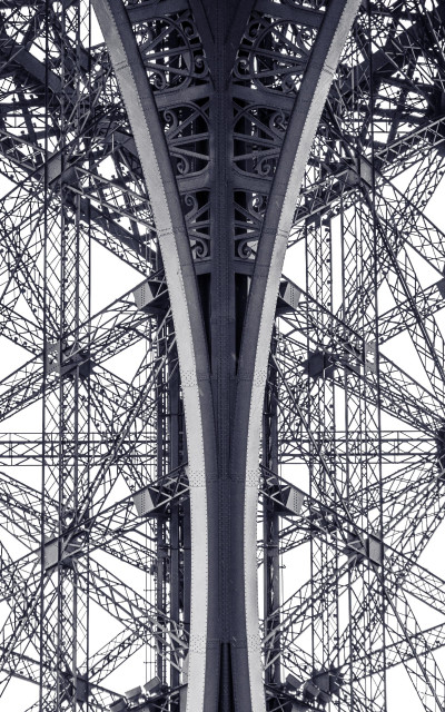 Monochrome photo taken from under the Eiffel Tower, looking straight up at the underside of one of the supporting arches and the surrounding ironwork structures.