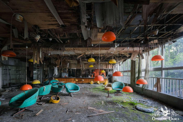 A decaying interior of an abandoned 1970s Japanese hotel with retro teal chairs, bright orange lamps, and mossy debris.