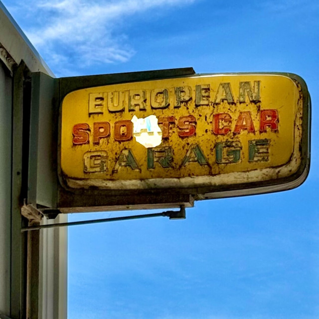 Sign for a place called European Sports Car Garage. It has been slowly deteriorating every time I've walked by. At this point, the R in sports seems to have been hit with a projectile. It's daytime, so you can see the A in car on the other side through the hole in the sign.