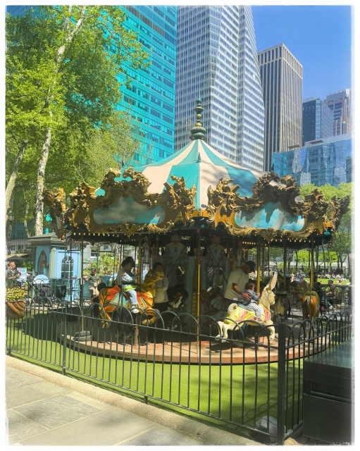 A color photo of a small carousel in Bryant Park. The bottom left hand corner shows some of the pavement and then there is a black rod iron fence around a big square piece of greenery. In the middle of that is an old fashioned carousel. It has a blue and white striped top with a lot of gold ornamentation around the edges. You can see people in the shadows standing and holding their young toddlers on the carousel horses. In the background is a large green tree on the left and several big office towers behind everything in different shades of blue. It shows a beautiful spring morning. 
