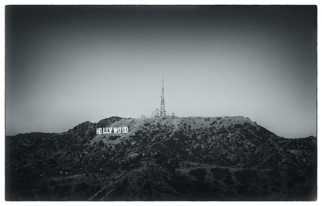A grainy, moody, faded, bluish-toned, monochrome view of the Hollywood sign and nearby transmission towers, on the crest of Mount Lee in the Hollywood hills. Taken from Griffith Park in March 2026.