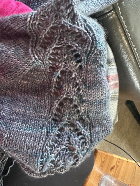 A close-up of a partially knitted item in variegated yarn, featuring intricate lace patterns.