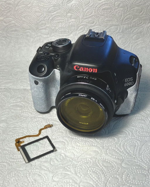 A photo of a canon eos 600d with a yellow coloured filter on its lens. A glass plane with a flatflex cable lays next to the camera 