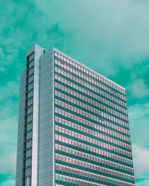 A photo up a weirdly iridescent office building. A few windows are open and reflect weirdly reddish. The sky is deeep vibrant turquoise 