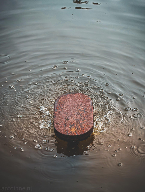 At the center sits a rust-colored, pill-shaped metal object, partially submerged in water. The texture is heavily weathered, showing deep oranges and browns.

At the point where the metal meets the surface, there is a ring of white foam and distinct air bubbles. Concentric, circular ripples radiate outward from the center, distorting the reflection of the sky.