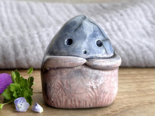 a vaguely dome-shaped nature spirit ceramic sculpture. it is glazed in a bottom-to-top gradient of soft reddish pink to blue and has plant and flower carvings on its body. it also has a gentle face, and two little arms across its front. it's standing on a piece of chestnut wood with grey muslin behind. next to it on the left are some speedwell — green leaves and tiny indigo-violet and white flowers — and a purple rock cress flower.