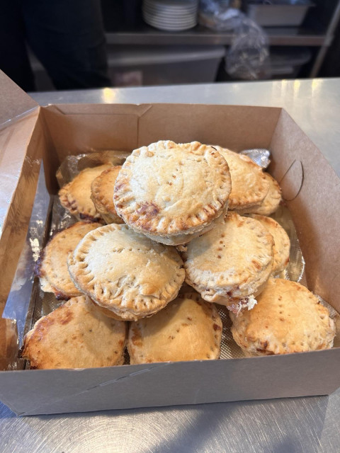 A tray of small pies sit in a cardboard takeout container. They are golden with fork decorations.

They are piled into a pie-ramid, 9, 4, 1 configuration.

The setting is a metal counter of a commercial kitchen with some plates and other things on low shelves in the far background.