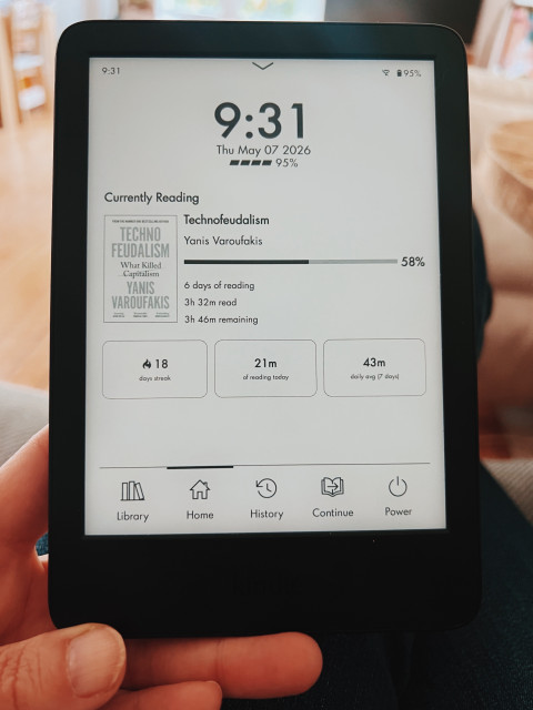 A photo of a hand holding a Kindle e-reader. The screen displays the SimpleUI interface with the hour on top (9:31), date, battery stats… and below a widget about the book I’m currently reading (Technofeudalism, I’m 58% in) and below another widget with reading stats: 18 days streak (with a flame symbol), 21 minutes of reading time today and 43 minute daily average over the past 7 days