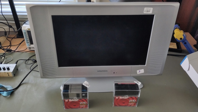 A small grey LCD TV and two transparent boxes of 3.5" floppy disks on a desk. 