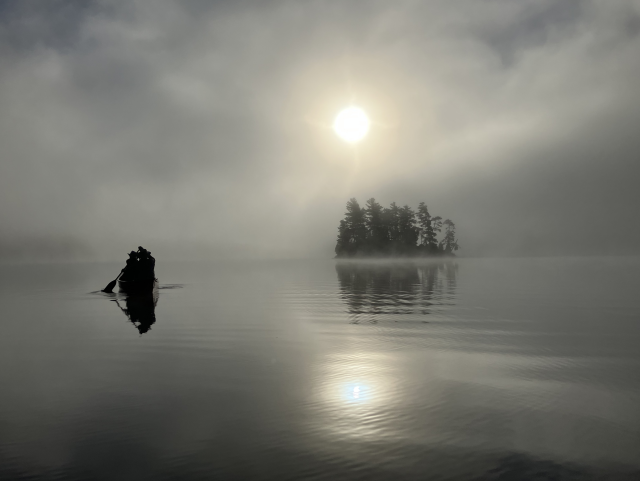 Looking towards the sun across a calm lake with the silhouette of an island in the distance on  the right, and a the silhouette of a canoe with two paddlers paddling into the distance on the left. The sun is behind rising fog.