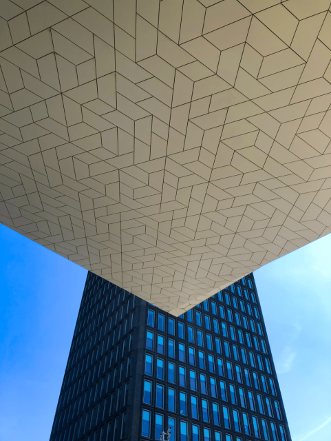 Shot looking at an exterior geometrically patterned surface of the Eye Filmmuseum in Amsterdam. Taken from a low angle angle, the image shows the surface occupying the top half of the frame with a triangular look. Beneath and in the background, against a clear blue sky, is a very tall modern building (flats? offices?) with many windows.