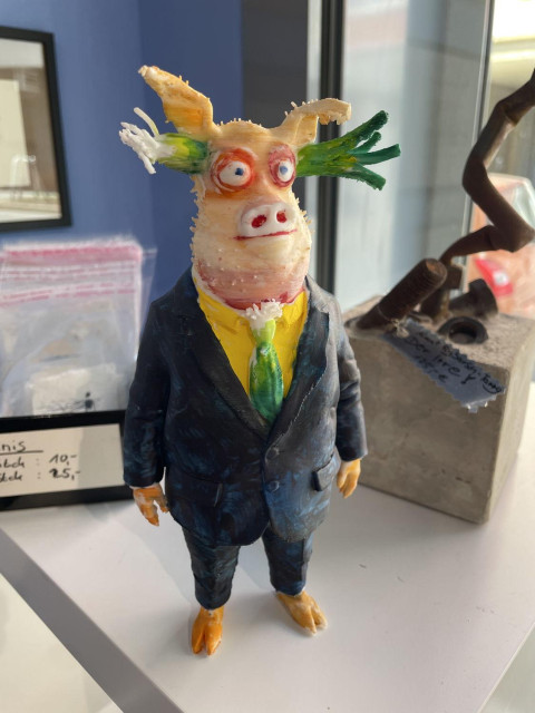 ein 3D-Druck einer figur, die Günter Rückert häufiger zeichnet - ein Schwein im Anzug mit grüner Krawatte und gelbem Hemd zu dunkelblauem Jackett. Die Figur hat eine Stange Lauch in den Ohren stecken - es sieht so aus, als sei diese ein mal quer durch den Kopf geschoben worden. Die Figur zeigt ein selbstzufriedenes Lächeln, hat rot umrandete Augen und einen Silberblick