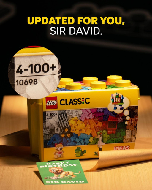 The image features a bright yellow LEGO Classic building bricks box prominently placed on a wooden surface. The box is filled with an assortment of colorful LEGO bricks, including red, blue, green, yellow, and pink pieces. The front of the box displays the LEGO Classic logo, followed by the text "CLASSIC" and a small bubble graphic showing a LEGO figure. Below this, there is text indicating "4-100+" and "10698", which is also highlighted in a circular inset in the upper left of the image. The phrase "IDEAS" is visible on the lower right of the box. In front of the LEGO box, there is a green "HAPPY BIRTHDAY" card featuring an illustration of a lion. The text "SIR DAVID" is printed on the card. A roll of brown craft paper is partially visible in the foreground, adding to a gift-wrapping theme. The background is dark and out of focus, with a yellow text overlay at the top that reads "UPDATED FOR YOU, SIR DAVID." The lighting is warm and highlights the vibrant colors of the LEGOs and the card. The composition is centered on the LEGO box and the birthday card, suggesting a gift presentation.