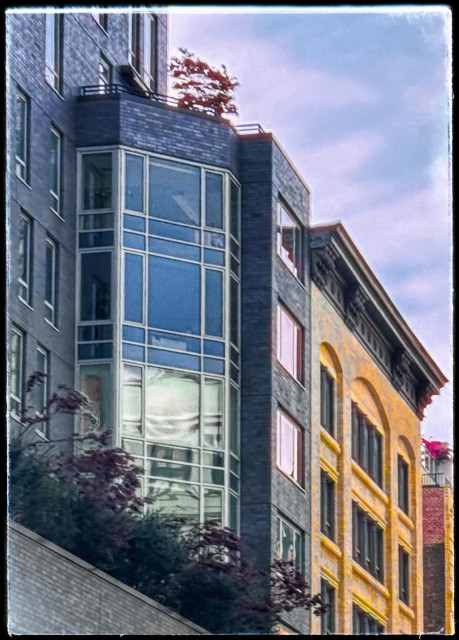 A color photo taken on a vertical angle of the windows of three different buildings. The the upper left is a flat gray brick building with a few windows and then sticking out at an angle is a set of rectangular Bay Windows that would give those apartments nice rounded views. On the top of those two bay windows is a balcony with a tree sticking out with red leaves. Next to that is a thin dark building with four square windows going down vertically that look light pink from the sun. Going down vertically the building next to that has a large dark balustrade on the top and is made of orange/yellow bricks in the shape of Arches with dark windows inside the arches. Next to it can be seen the sliver of a building with something red sitting on top in the distance. The upper right hand corner is light blue sky with wispy pink clouds. The triangle in the bottom left-hand corner is the top of a white brick balcony that has dark green leaves and branches with purple leaves overhanging it. 
