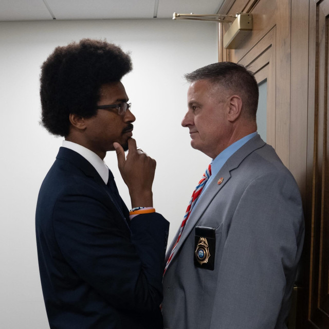 Tennessee State Representative Justin Pearson, a black man with a two-inch afro and wearing a dark blue suit, stands with his right hand raised to his chin in a contemplative pose. He stands chest-to-chest with a white security guard with a brush cut wearing a grey suit on which a badge hangs from the breast pocket, and who is blocking the door to the legislative chamber.