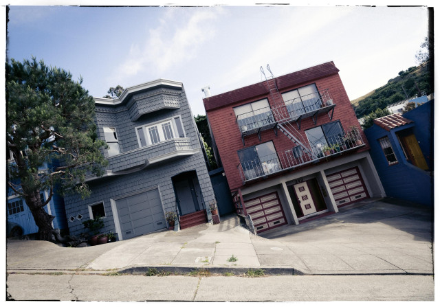 A picture of two houses on a very steep section of Ripley Street in the Bernal Heights neighborhood of San Francisco. Photographed with the camera at an angle that keeps the street level rather than the houses, these two 2 and 3-story homes appear to lean drunkenly to the left. It's Friday so that's understandable. TGIF and all.