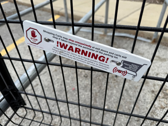 A close-up shot of a white warning sign attached with screws to the black wire mesh of a shopping cart. The sign features a red stop hand icon on the left and a shopping cart icon with signal waves on the right. Bold red and black text reads: "WARNING! Shopping Carts may stop unexpectedly at exit doors and carts will stop if taken beyond the perimeter of the parking lot." The message is also provided in Spanish below. The background shows a blurry grey asphalt parking lot and yellow lines.