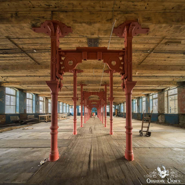Wide-angle view of an abandoned industrial building with rows of red cast iron columns supporting a wooden ceiling; debris and old equipment are scattered, and light streams through large windows.