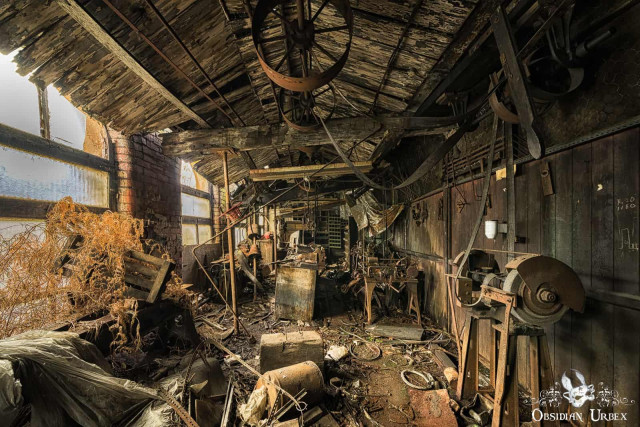 A heavily decayed, abandoned Victorian textile mill workshop features rusty machinery, scattered debris, and exposed wooden beams. Sunlight enters through broken windows.