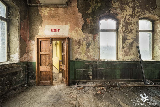 An abandoned room within a decaying Victorian textile mill. Peeling walls, three windows, and a prominent wooden door marked "EXIT" are visible.