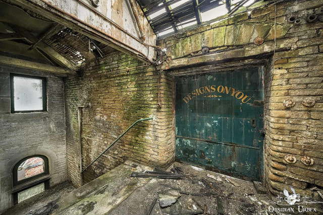 Placeholder Inside an abandoned Victorian mill, moss-covered brick walls frame a decaying room. A prominent green door reads "DESIGNS ON YOU," with debris scattered on the floor and a crumbling roof above.