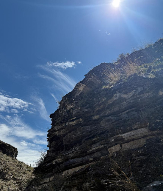 cliff composed of layered rocks have wisps of yellow grass which catch the bright sunlight and mirror the wispy clouds in the blue sky
