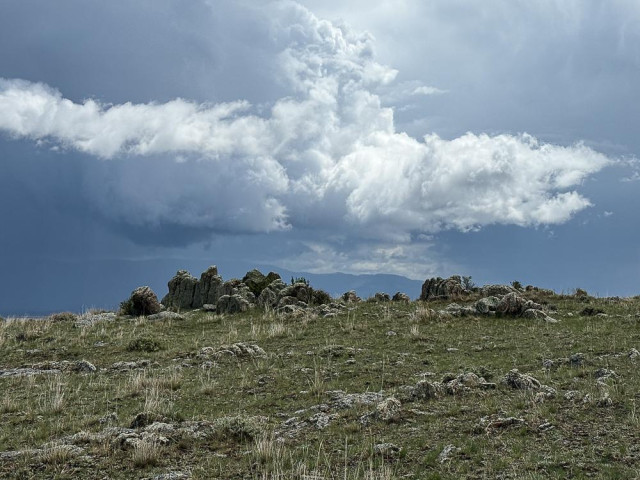 an odd shaped white cloud hovers over a similarly shaped rock formation on a hill. The sky behind is dark with rainclouds