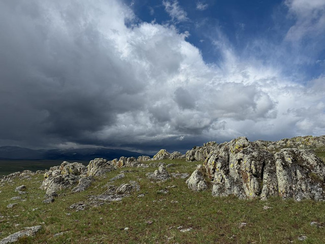 white puffy clouds and wispy icy clouds mingle above a hillside of grass and lichen-covered rocks
