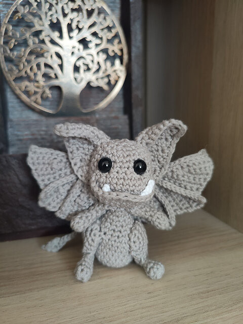 A small crocheted creature made from light gray yarn sits on a wooden surface. It has a round head with two pointed ears, large black safety eyes, and two short white fangs. Its body is squat with short limbs, and a pair of wide, slightly curved wings extends from its back. Behind the figure is a decorative metal panel featuring a cut‑out tree design, softly lit from behind.
