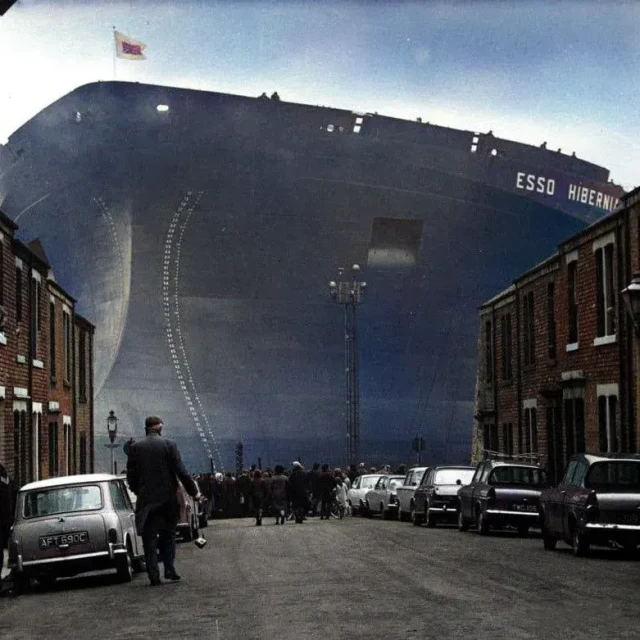An imposing supertanker towers over a red brick street with cars parked on both sides. A man in a hat walks towards the ship. The size of the ship is perhaps an effect of a photoscopic lense zooming in from a distance