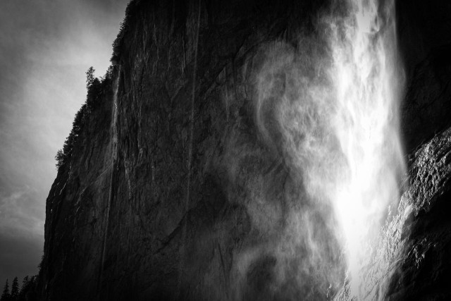 Schwarzweissbild von einer dunklen Felswand von der ein Wasserfall stürzt. Die Sonne scheint in die Gischt und lässt ihn hell leuchten.