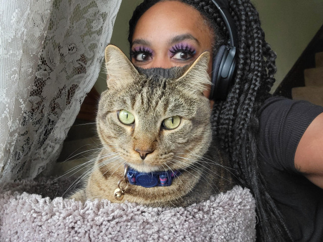 An adorable tabby cat sits in a perch with a woman smiling behind him though only her eyes are visible 