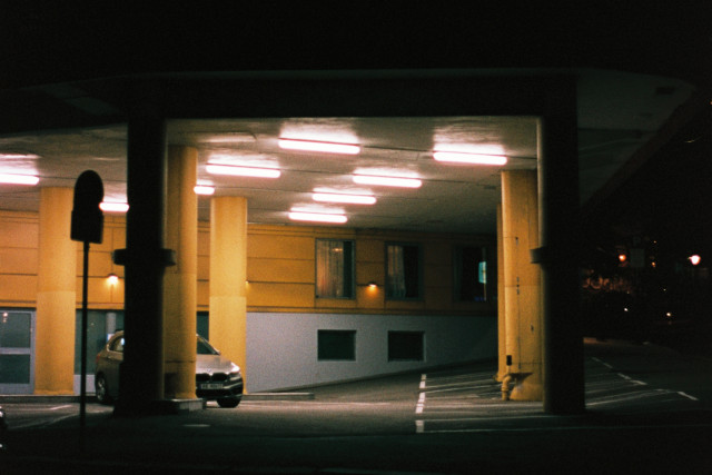 Night-time photo of a parking lot under a roof with lots of glowy, fluorescent light tubes. The yellow concrete pillars holding up the roof match the yellow wall in the back. A single car is attempting to hide behind one of the pillars.