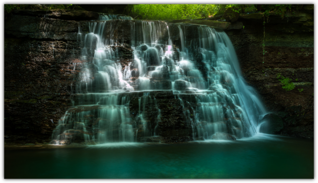 Ein mehrstufiger Wasserfall, der über dunkle Felsen in ein ruhiges türkisfarbenes Becken fließt, mit weichem, lang belichtetem Wasser vor einem grünen, sonnigen Hintergrund.