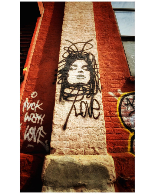 Low-angle photo looking up at the red brick facade of a building. A vertical section protrudes slightly and is painted a lighter color than the warm red of the rest of the facade. On this is displayed a graffiti image of an upturned face with eyes closed, framed by black swirls and squiggles suggesting hair, and with the word LOVE underneath. To the left is a message in white spraypaint: FUCK WITH LOVE. Another bit of graffiti can be partially seen at right, in outlines of black, yellow, and white. Taken on Van Brunt Street in Red Hook, Brooklyn.