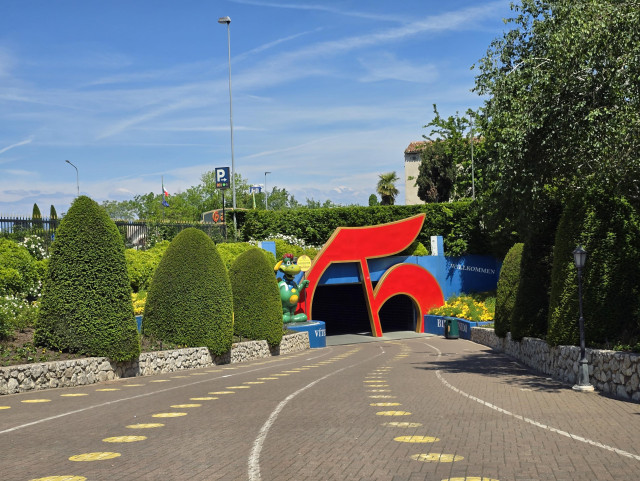 A sunny outdoor entrance to a theme park, with a large red G-shaped structure, manicured hedges, flowers, a cartoon dragon figure, and a pathway leading into a tunnel.