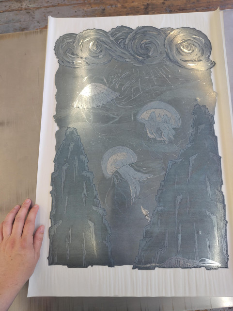 The whole etching plate with hand for scale. The plate is a sheet of zinc with a laser cut outer edge that follows the contours of the rocks and waves