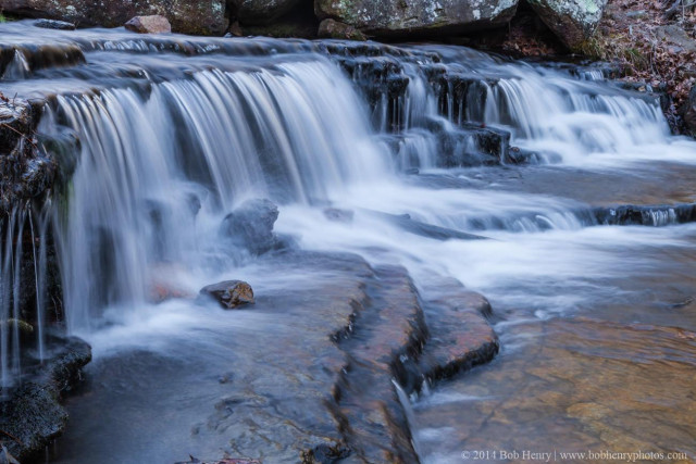 A horizontal image of a wide, tiered waterfall cascading over dark, mossy rock ledges into a shallow pool. The long exposure creates a soft, silky white veil effect against the rugged stone.