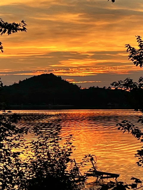 Red and yellow sunset with subtle dark striations in the clouds giving definitions to the layers of clouds. Silhouettes of tree branches frame the picture and a silhouette of a sand dune is at the far side of the lake. The colors are reflected off the lake with checkerboard ripples.