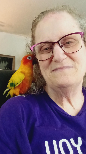 Me, looking straight ahead. I am smiling, my hair is pulled back and I am wearing purple prescription glasses. My sun conure parrot, Sherbert, is sitting on my shoulder with her beak pressed against my cheek. She is about 12 inches from beak to tail, is mostly yellow and orange, has some green and blue on her wings and tail, she has dark grey eyes and a black beak.