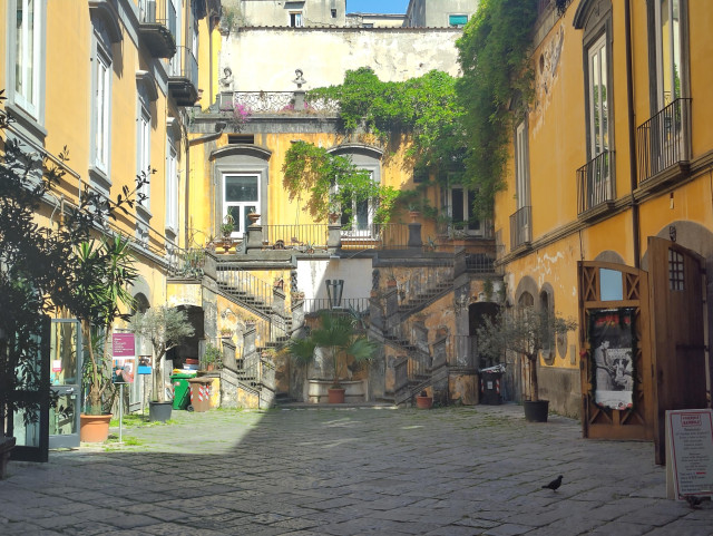 Photo of a courtyard in Naples. It's yellow and a shadow cuts diagonal across the back wall. Plants, walls and stairs cases are all around.
