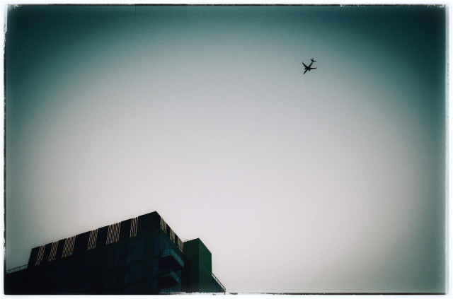 A grainy, greenish, heavily vignetted photo of a plane flying through the skies high above the iceberg tip of a skyscraper in downtown Los Angeles. Kind of artsy. Minimalist.