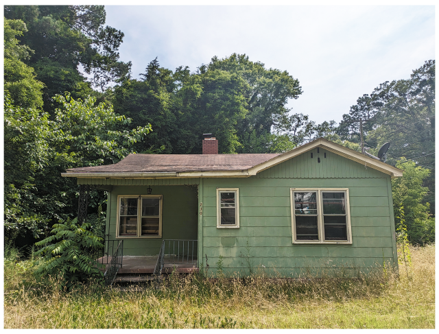 	
a  small, weathered green house with a front porch, overgrown grass, and bushes sits in front of dense trees. the roof is brown with a central chimney, and the siding shows signs of age. Three front windows and one porch light are visible.